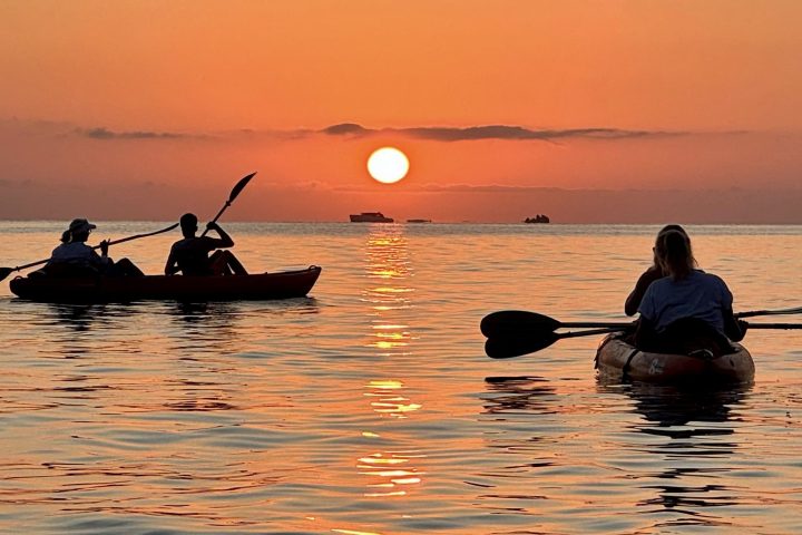 Two kayaks on calm water at sunset with an orange sky and visible sun.
