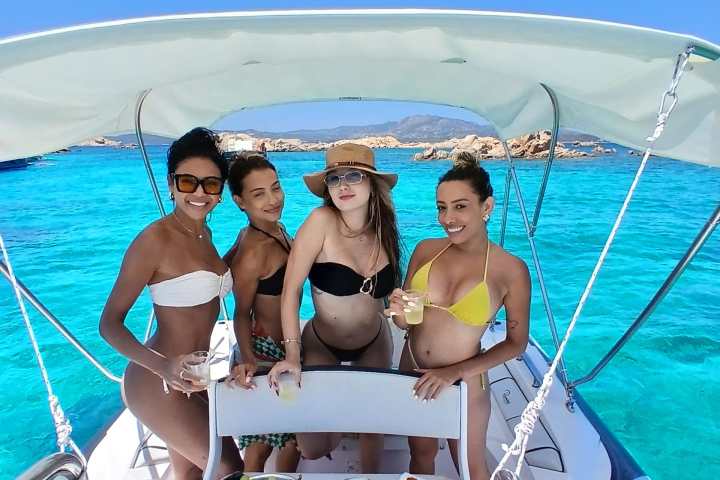 Four women in swimwear on a boat with snacks and drinks, surrounded by clear blue water.
