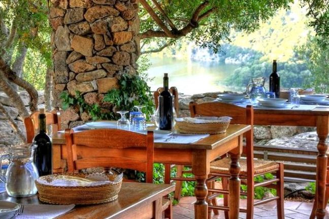 Outdoor dining area with wooden tables, chairs, and a stone wall surrounded by trees.
