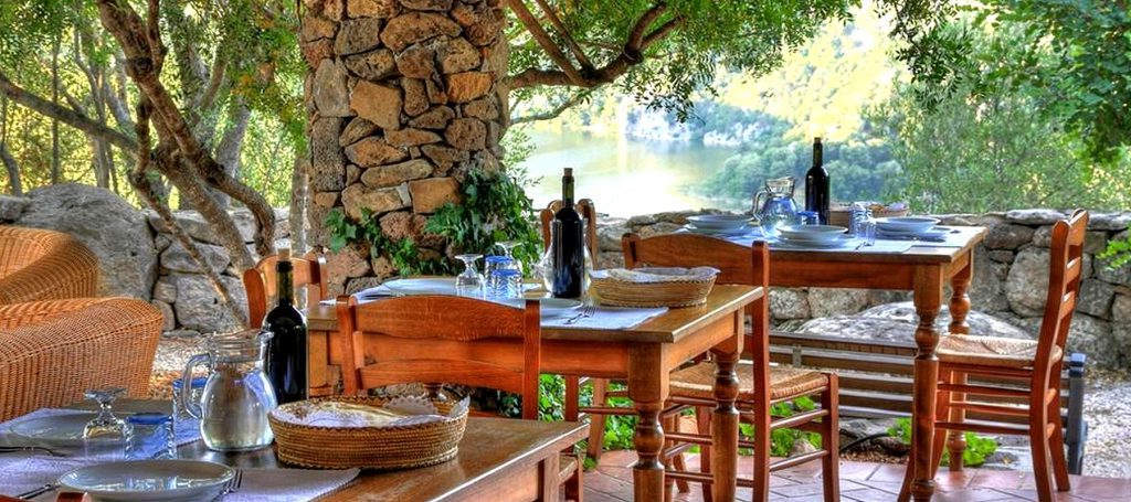 Outdoor dining area with wooden tables, chairs, and a stone wall surrounded by trees.