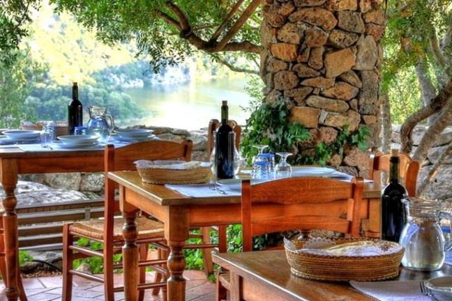 Outdoor dining area with wooden tables, chairs, and a stone wall surrounded by trees.