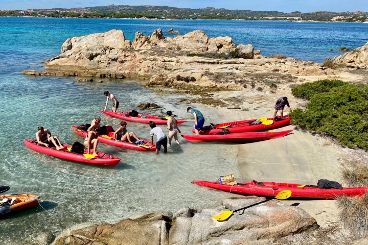 People with red kayaks gather on a rocky shore with clear blue water and distant hills.