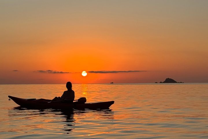 Silhouetted kayaker on calm sea at sunset, with distant islands and an orange sky.
