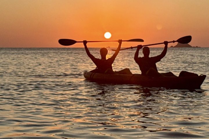Silhouetted kayakers at sunset on calm sea, holding paddles overhead.