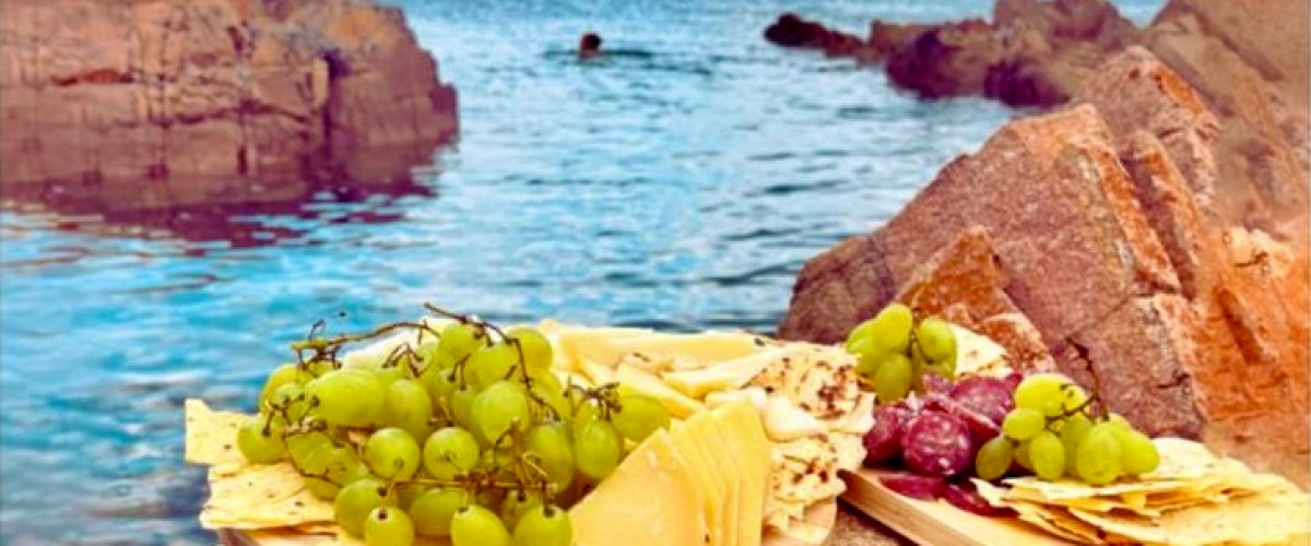 Cheese and grape platter on rocks by the sea with a swimmer in the water.