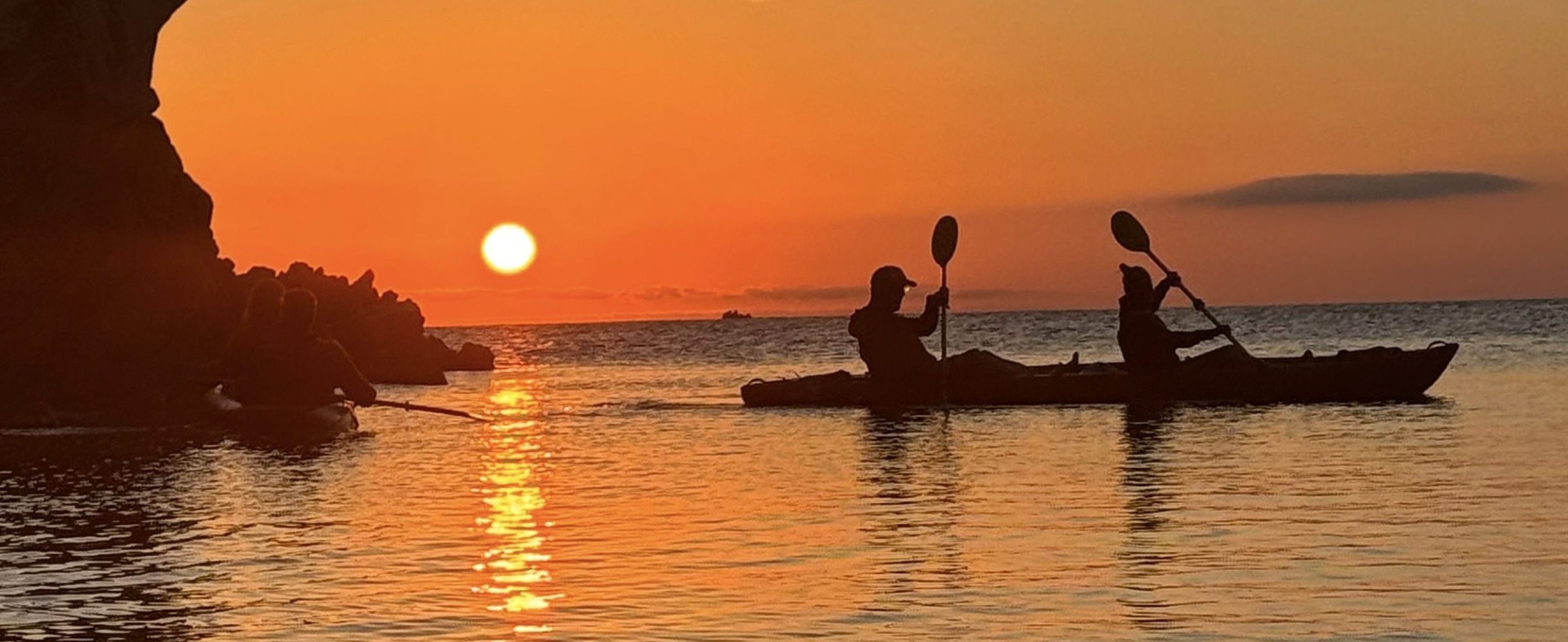 Two kayakers silhouetted against a sunset over calm water near rocky cliffs.