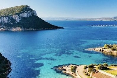 Coastal landscape with blue sea, small beach, green trees, and a rocky hill in the distance.